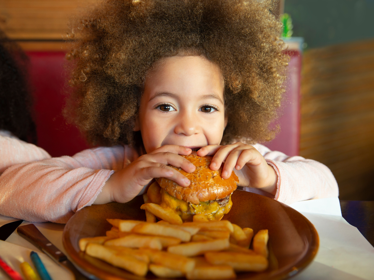kids eat free restaurants kids eat free restaurants: A young child with curly hair enjoying a burger at a restaurant, with a plate of fries in front of them. The child is smiling and holding the burger with both hands
