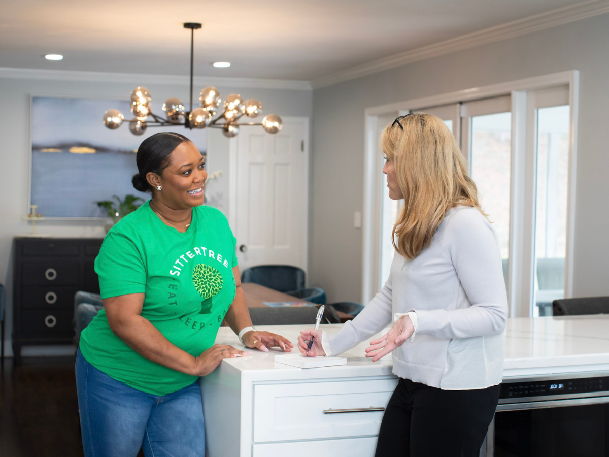 Preparing for a Babysitting Job Preparing for a Babysitting Job: A babysitter wearing a green SitterTree shirt stands in a kitchen, smiling and having a conversation with a parent holding a pen and notebook, discussing job details