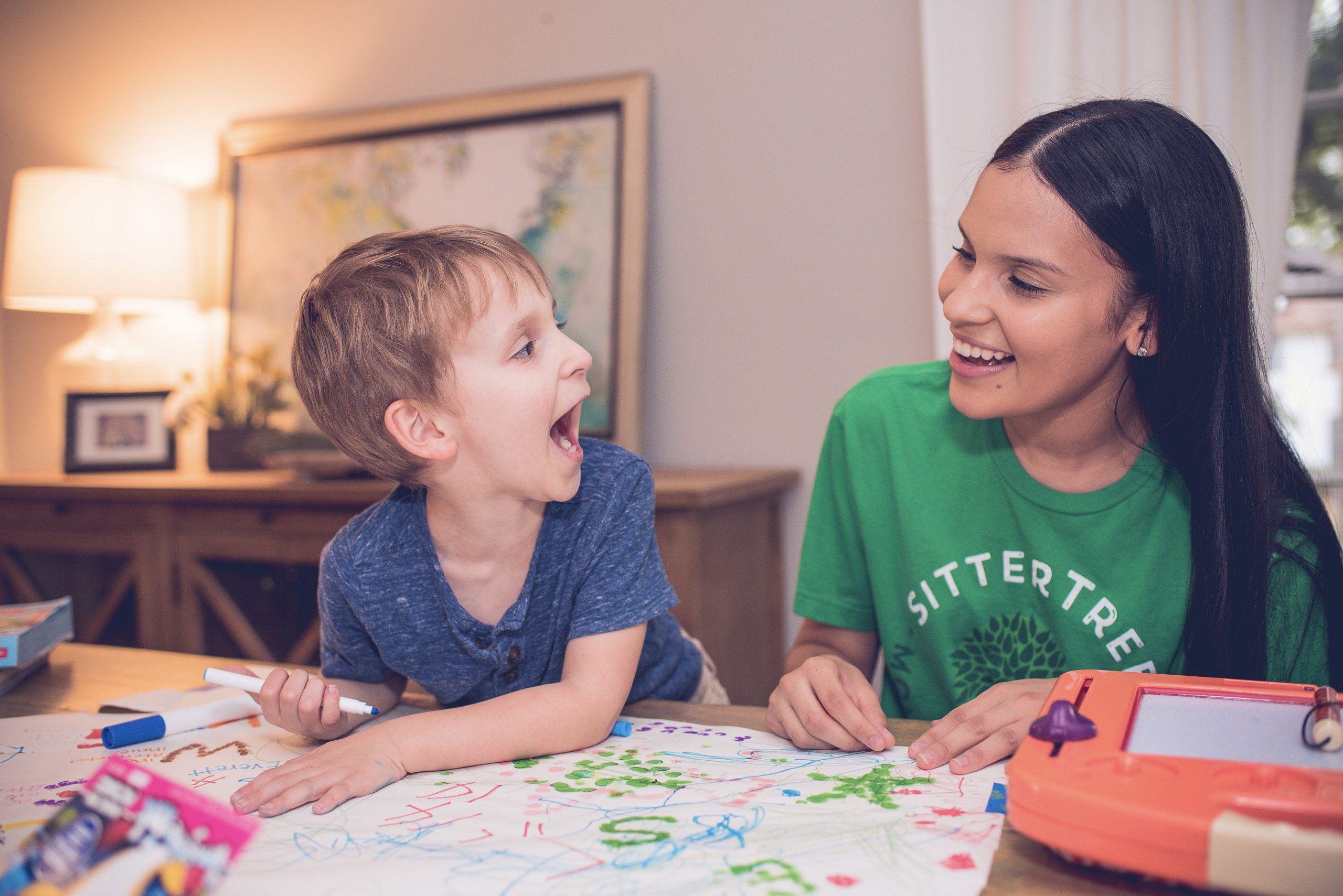 Creative play ideas for school-aged children A babysitter wearing a green SitterTree t-shirt laughs with a young boy as they draw together on a colorful piece of paper in a cozy room: Creative play ideas for school-aged children