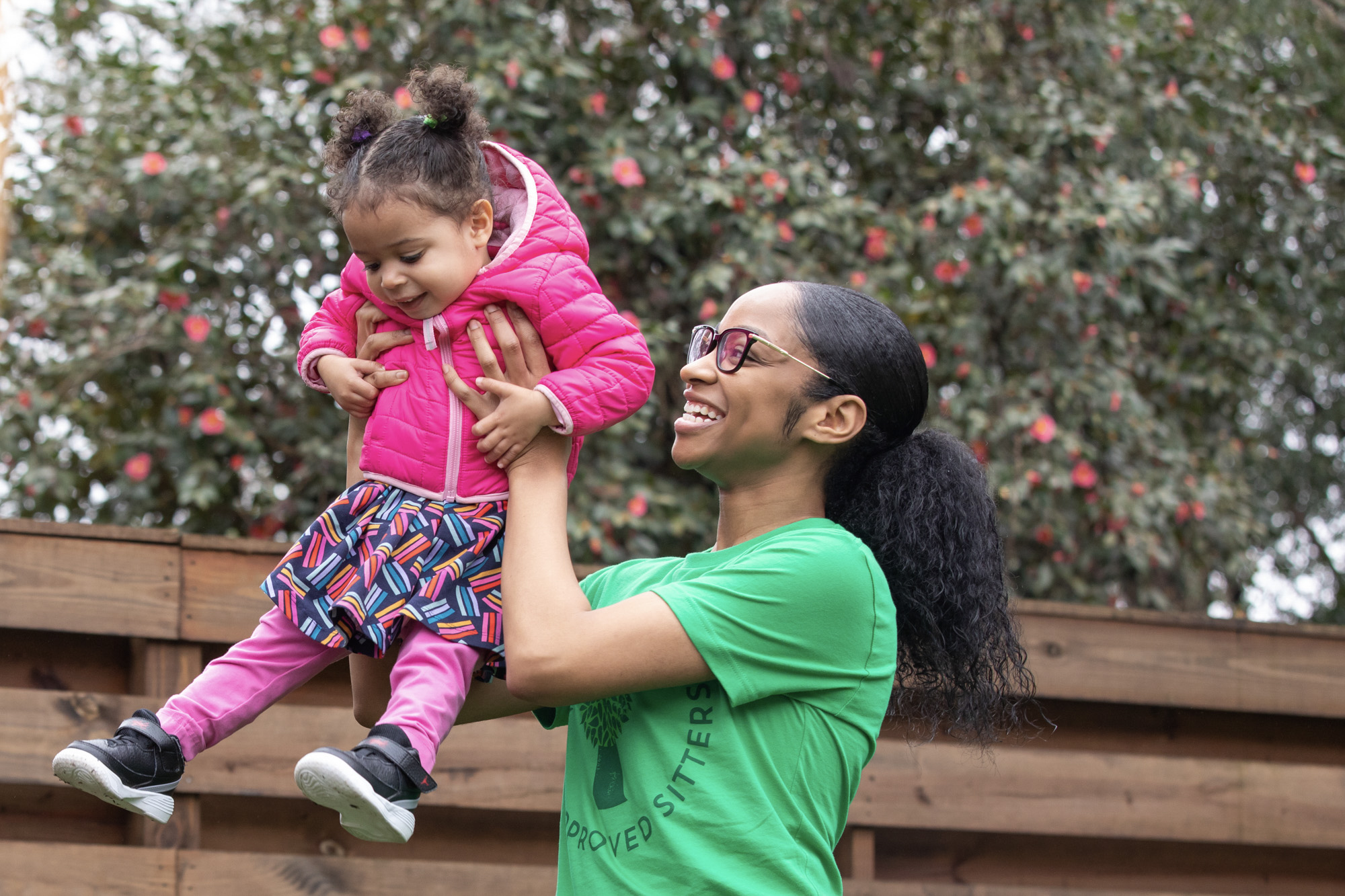 A babysitter wearing a green SitterTree t-shirt lifts a smiling toddler in a pink jacket and colorful skirt into the air, with a wooden fence and blooming tree in the background: How to find a babysitter near me